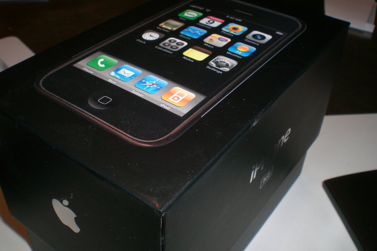 The original 2007 iPhone resting on top of its black retail box with the home screen lit up.