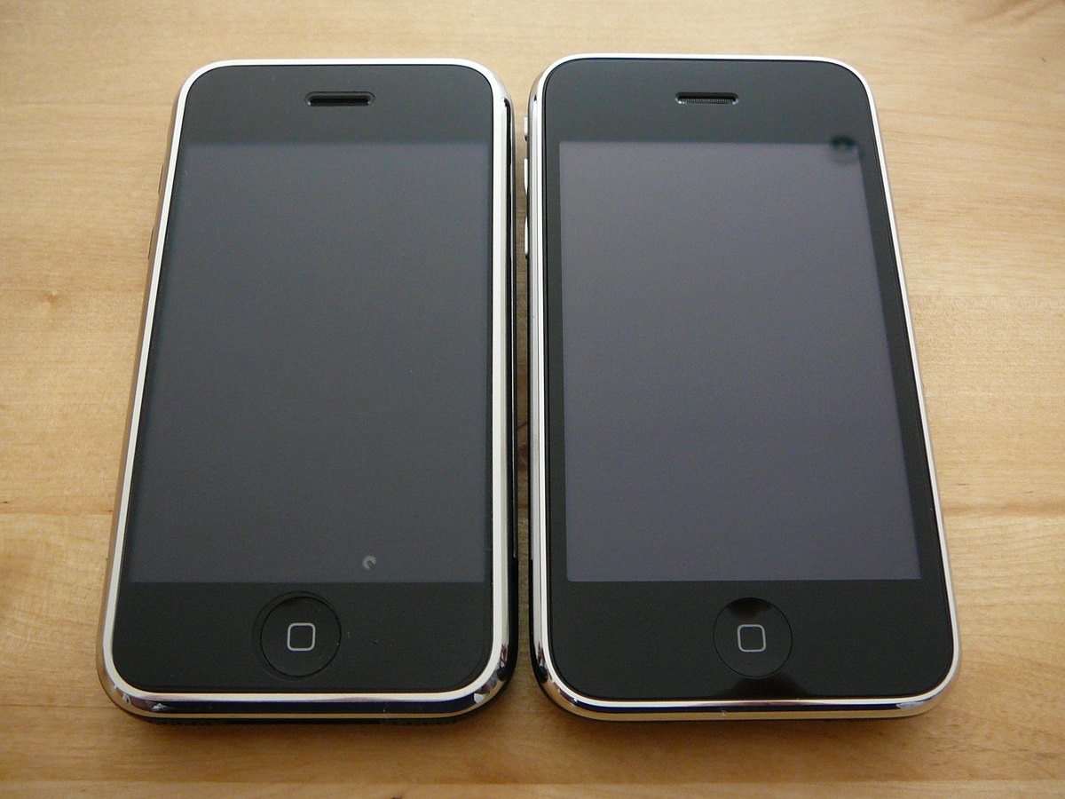 The original 2007 iPhone on the left next to the slightly larger iPhone 3G on the right, both face up on a wooden table showing their identical home button and bezel design.