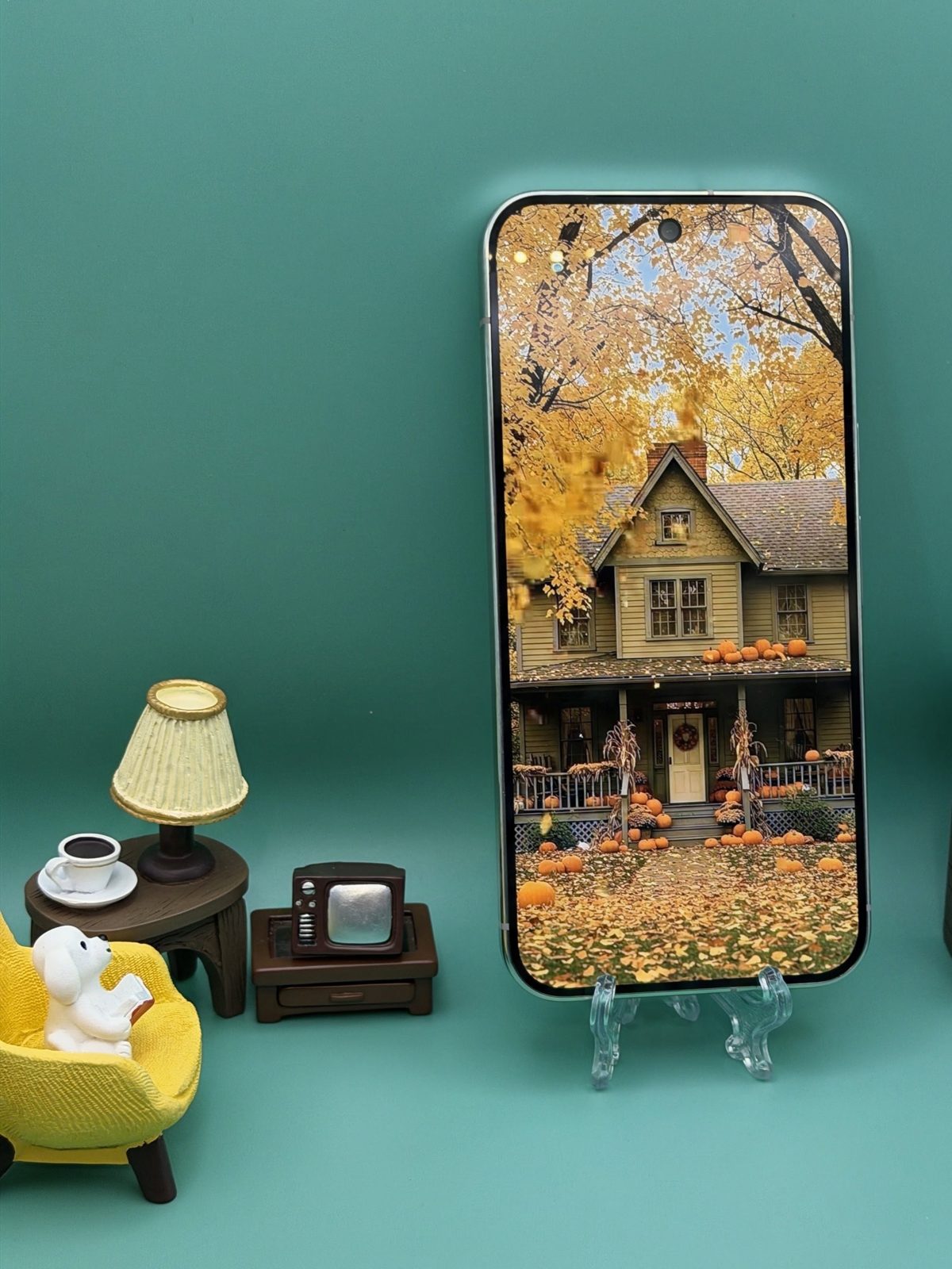 A Pixel 11 Pro XL standing on a clear easel showing a full screen autumn photograph of a yellow house surrounded by pumpkins and fallen leaves, with miniature furniture beside it against a teal background.