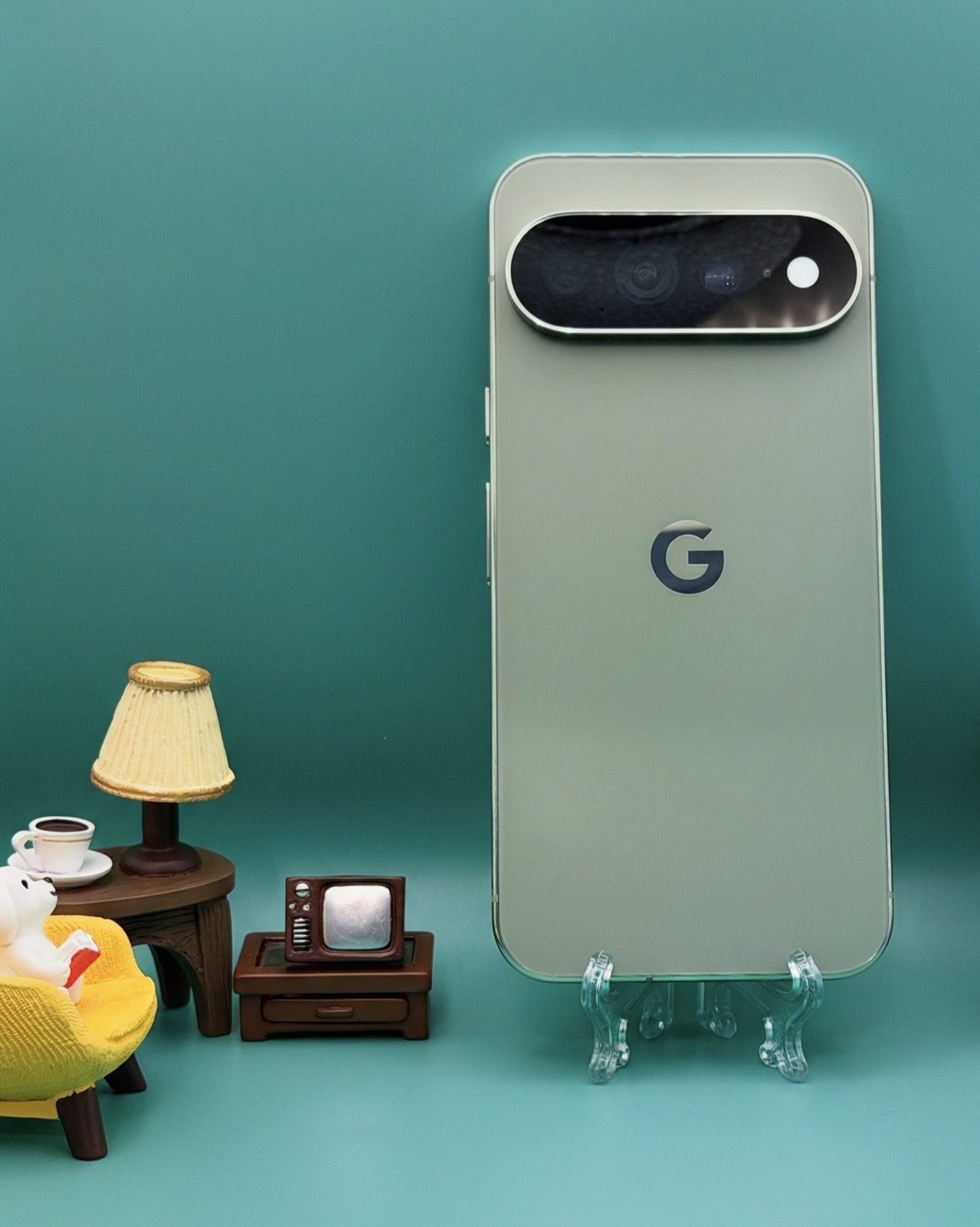 The back of a Pixel 11 Pro XL in a pale sage finish on a clear easel against a teal background, its oval camera bar visible, with miniature furniture beside it.