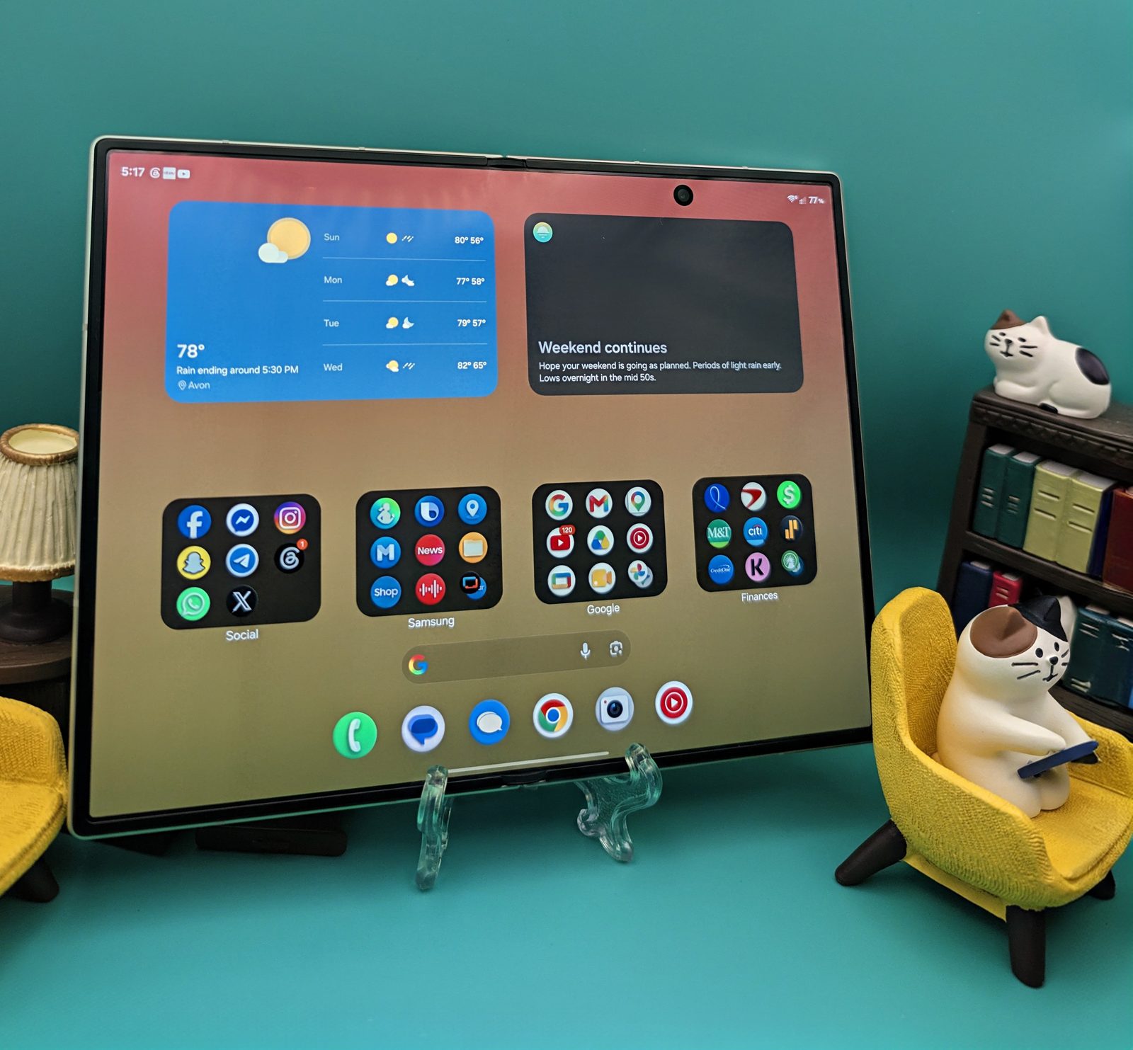 A Galaxy Z Fold 8 unfolded and standing on a clear easel, its wide inner display showing a weather widget and four app folders, with miniature furniture and a small bookshelf arranged around it against a teal background.