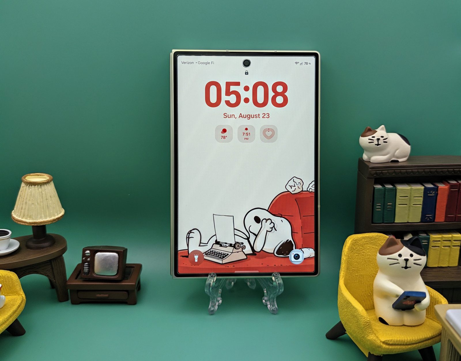A Galaxy Z Fold 8 closed and standing upright on a clear easel, its wide cover screen showing a lock screen with a large clock, miniature furniture arranged around it.